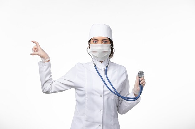 Vorderansicht Ärztin im weißen sterilen medizinischen Anzug und mit Maske aufgrund von Covid-Stethoskop auf White-Wall-Disease-Virus-Covid-Pandemie-Krankheit