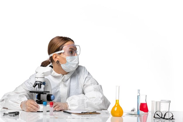 Vorderansicht Ärztin in Spezialanzug und Maske unter Verwendung eines Mikroskops auf weißem Hintergrund Covid-Virus-Pandemie-Coronavirus