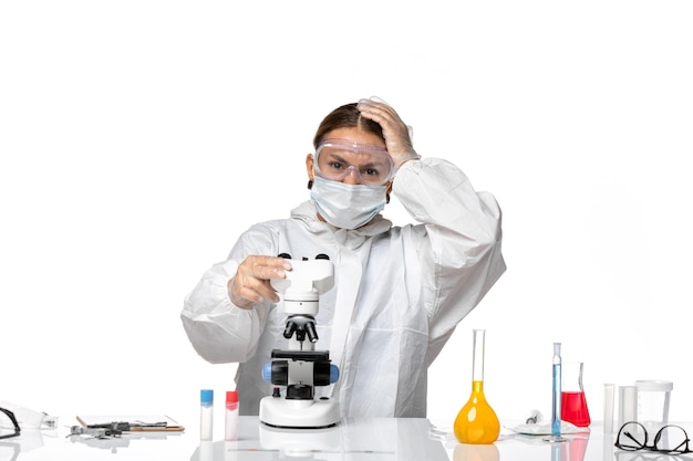Vorderansicht Ärztin in Spezialanzug und tragende Maske unter Verwendung eines Mikroskops auf weißem Hintergrundvirus-Covid-Pandemie-Coronavirus