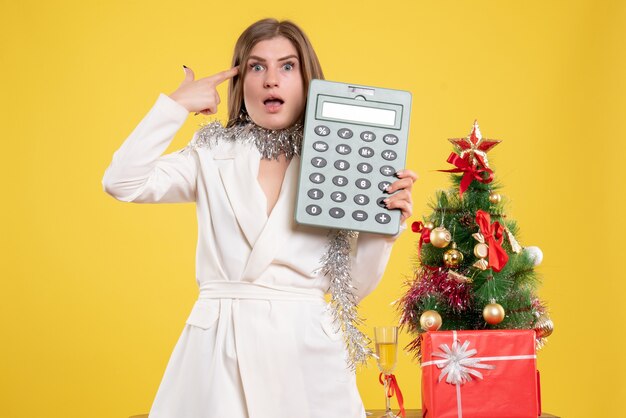 Vorderansicht Ärztin stehend und hält Rechner auf gelbem Hintergrund mit Weihnachtsbaum und Geschenkboxen