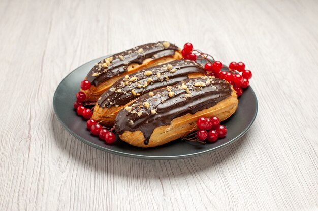 Vorderansicht Schokoladen-Eclairs und Johannisbeeren auf der grauen Platte auf dem weißen hölzernen Hintergrund