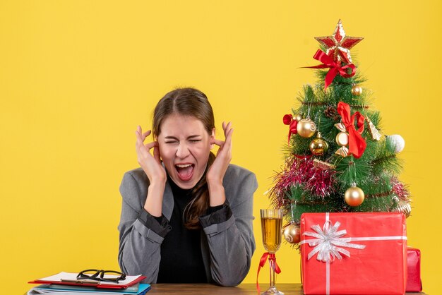 Vorderansicht schrie Mädchen, das am Schreibtisch sitzt Ohren mit ihren Fingern Weihnachtsbaum und Geschenkcocktail bedeckt
