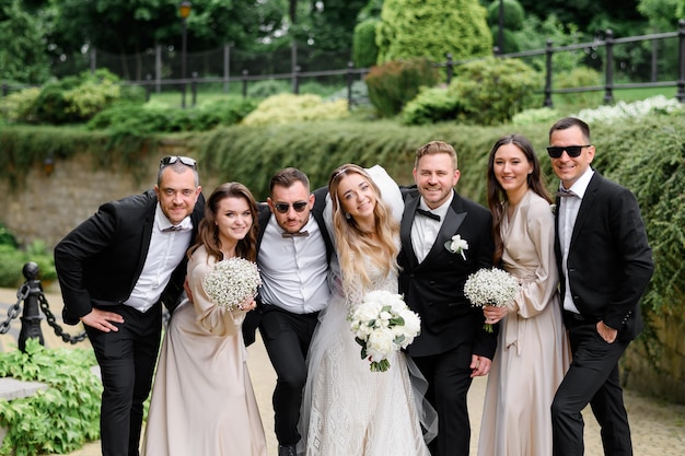 Vorderansicht stilvoller Freunde und Jungvermählten in Festival-Outfits, die sich lächelnd umarmen und während der Hochzeit auf Parkhintergrund in die Kamera posieren