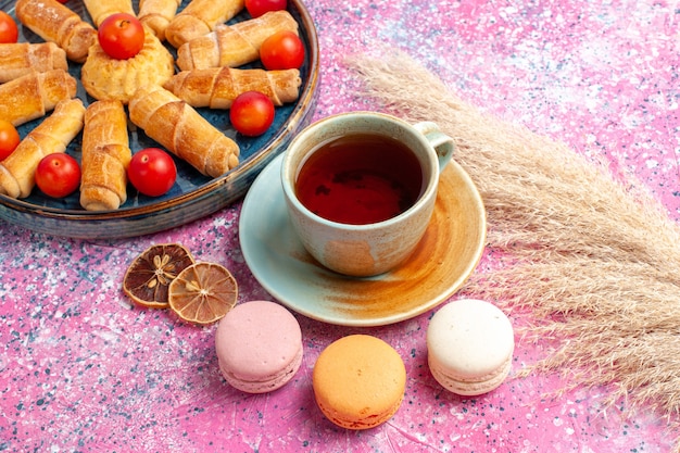 Vorderansicht süße köstliche Bagels in Tablett mit frischen sauren Pflaumen französischen Macarons und Tasse Tee auf hellrosa Schreibtisch