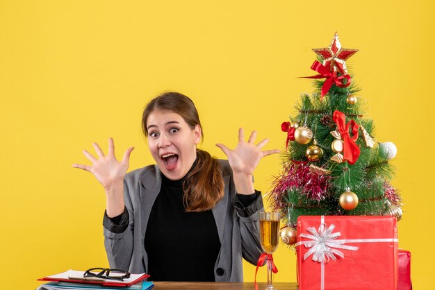 Vorderansicht überraschte glückliches Mädchen, das am Schreibtisch mit geöffnetem Handweihnachtsbaum und Geschenkcocktail sitzt