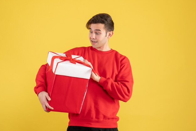 Vorderansicht überraschte jungen Mann mit rotem Pullover, der Geschenk auf Gelb hält