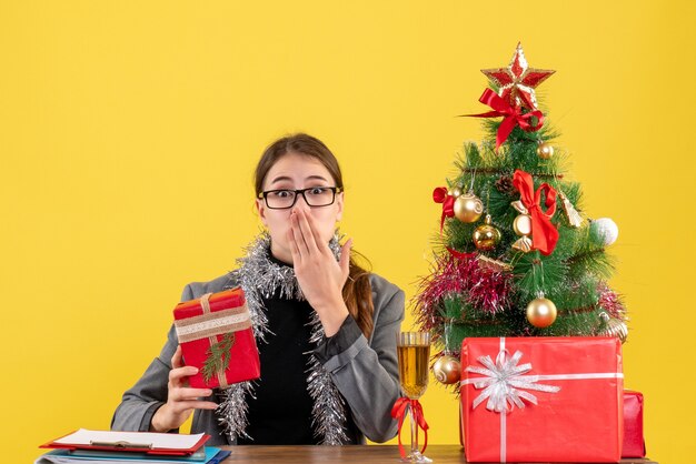 Vorderansicht überraschte Mädchen mit Brillen, die am Tisch sitzen und Hand an ihren Mundweihnachtsbaum und Geschenkcocktail setzen