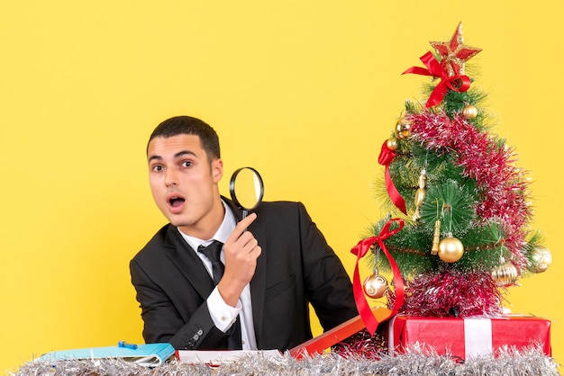 Vorderansicht überraschte Mann mit optischer Lupa in der Hand, die am Tischweihnachtsbaum und an den Geschenken sitzt