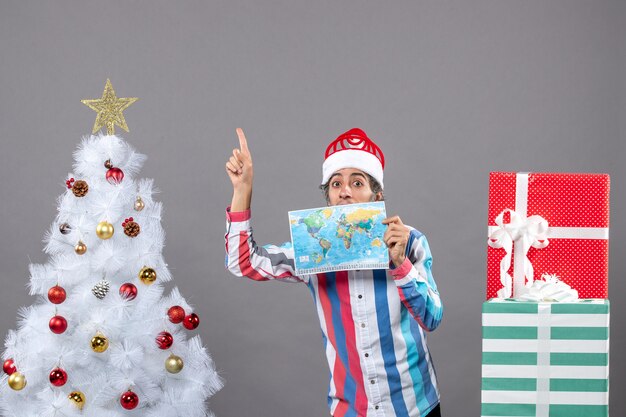 Vorderansicht überraschte Mann mit Spiralfeder Santa Hut, der Weltkarte hält, die auf einen hohen Platz zeigt
