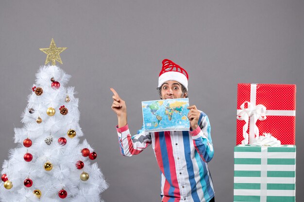 Vorderansicht überraschte Mann mit Spiralfeder Santa Hut, der Weltkarte hält, die auf Weihnachtsbaum zeigt