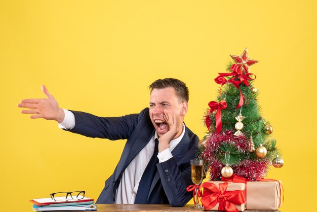 Vorderansicht verärgerter Mann, der schreit, während er am Tisch nahe Weihnachtsbaum und Geschenke auf gelbem Hintergrund sitzt