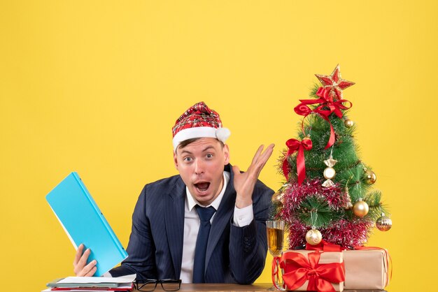 Vorderansicht verblüffter Geschäftsmann, der am Tisch nahe Weihnachtsbaum sitzt und auf gelbem Hintergrund präsentiert