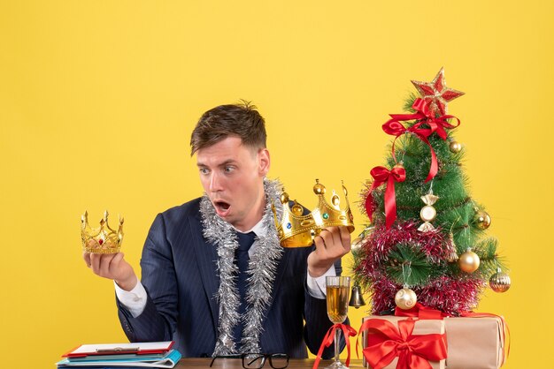 Vorderansicht verblüffter Geschäftsmann, der Kronen betrachtet, die am Tisch nahe Weihnachtsbaum sitzen und auf gelbem Hintergrund präsentieren