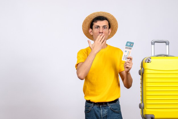 Vorderansicht verwirrte jungen Mann im gelben T-Shirt und im Strohhut, die nahe gelbem Koffer stehen