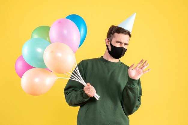 Vorderansicht verwirrte jungen Mann mit Partykappe und bunten Luftballons, die auf Gelb stehen