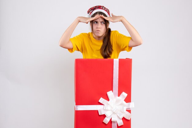 Vorderansicht verwirrte Mädchen mit Weihnachtsmütze, die Hände zu ihrer Stirn stellte, die hinter großem Weihnachtsgeschenk steht