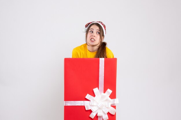 Vorderansicht verwirrtes Mädchen mit Weihnachtsmütze, die hinter großem Weihnachtsgeschenk steht