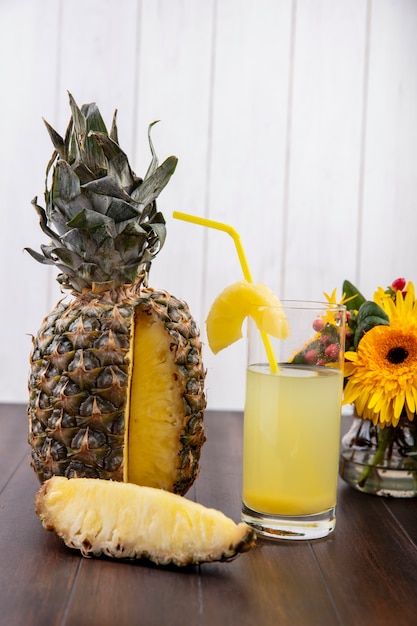 Vorderansicht von Ananas und Ananasscheibe und Saft im Glas mit Trinkrohr und Blumen auf Holzoberfläche