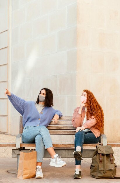 Vorderansicht von Freundinnen mit Gesichtsmasken draußen sitzen auf einer Bank mit Kopierraum