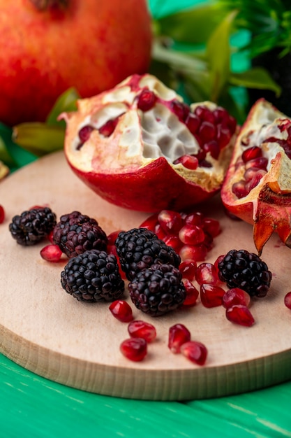 Vorderansicht von Früchten als Brombeer- und Granatapfelstücke und Beeren auf Schneidebrett mit ganzem Granatapfel und Blättern auf grüner Oberfläche