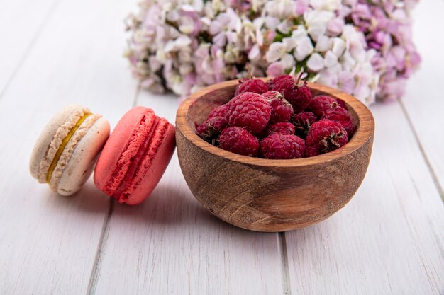 Vorderansicht von Himbeeren in einer Schüssel mit Macarons und einem Blumenstrauß auf einer weißen Oberfläche