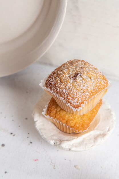 Vorderansicht von kleinen leckeren Kuchen in Papierformen mit Zuckerpulver auf der weißen Oberfläche