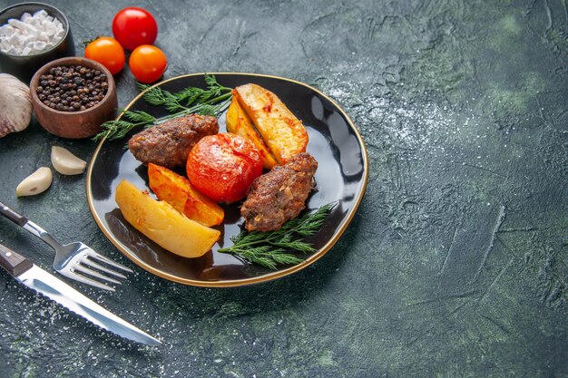 Vorderansicht von köstlichen Fleischkoteletts, die mit Kartoffeln und Tomaten auf einem schwarzen Tellerbesteck gebacken werden, Gewürze Knoblauchtomaten auf der rechten Seite