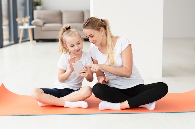 Vorderansicht von Mutter und Tochter zu Hause auf Yogamatte, die auf Smartphone spielt