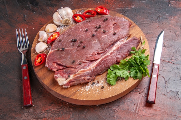 Vorderansicht von rotem Fleisch auf Holztablett und Knoblauch, grüner Zitronenpfeffer, Zwiebelgabel und Messer auf dunklem Hintergrund