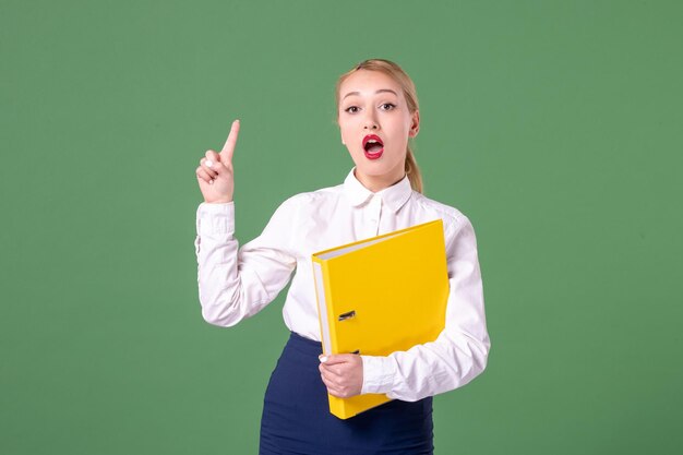 Vorderansicht weibliche Lehrerin, die in strenger Kleidung mit gelben Dateien auf grünem Hintergrund posiert