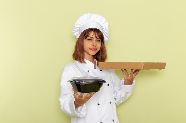 Vorderansicht weiblicher Koch im weißen Kochanzug, der Nahrungsmittelbox und schwarze Schüssel auf grüner Oberfläche hält
