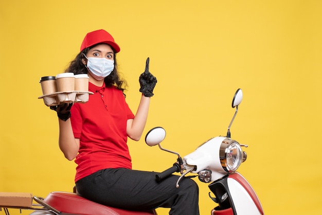 Vorderansicht weiblicher Kurier in Maske auf Fahrrad mit Kaffeetassen auf gelbem Hintergrund Arbeiterservice Pandemie Uniform Job Frau Lieferung Covid-