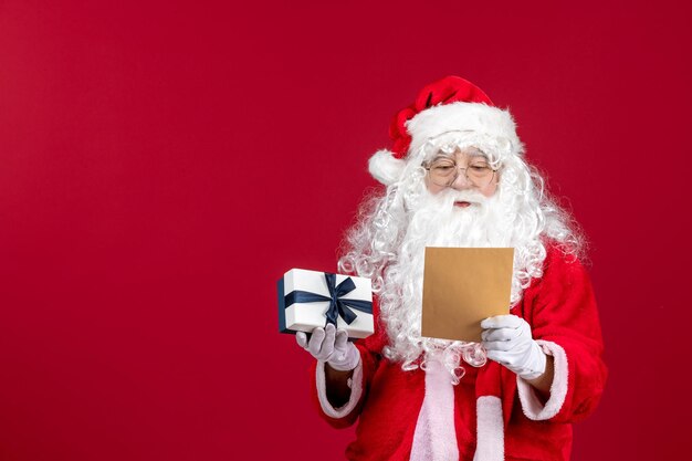 Vorderansicht Weihnachtsmann liest Brief vom Kind und hält Geschenk auf dem roten Emotionsgeschenk Weihnachtsfeiertag