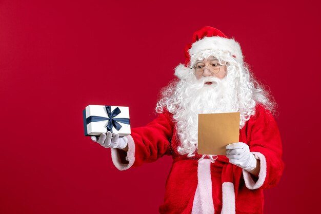 Vorderansicht Weihnachtsmann liest Brief vom Kind und hält Geschenk auf dem roten Emotionsgeschenk Weihnachtsfeiertag