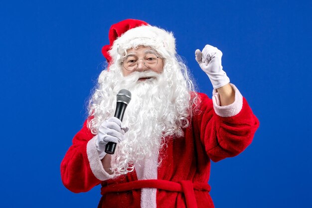 Vorderansicht Weihnachtsmann mit rotem Anzug und weißem Bart, der Mikrofon auf blauem Neujahrsfest hält