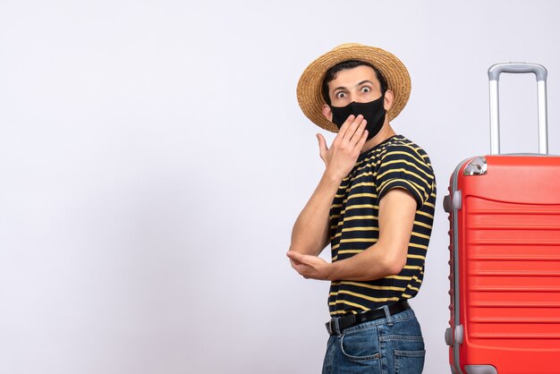 Vorderansicht wunderte sich junger Tourist mit schwarzer Maske, die in der Nähe des roten Koffers steht