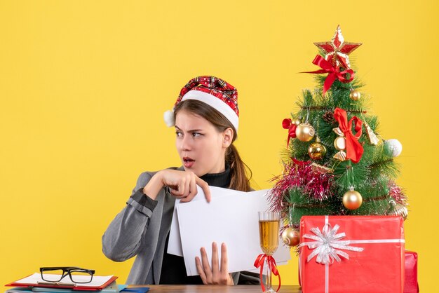 Vorderansicht wunderte sich Mädchen mit Weihnachtshut, das am Tisch sitzt, der Dokumentweihnachtsbaum und Geschenkcocktail hält