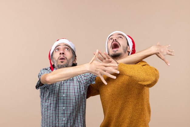 Vorderansicht zwei aufgeregte Kerle mit Weihnachtsmützen, die beide etwas vermeiden, während sie hoch auf beigem isoliertem Hintergrund schauen