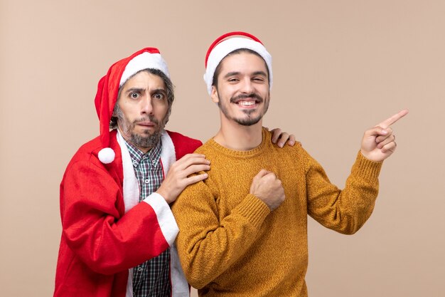 Vorderansicht zwei Freunde mit Weihnachtsmützen, die eine Freundschulter halten und die andere Richtung auf beige lokalisiertem Hintergrund zeigen