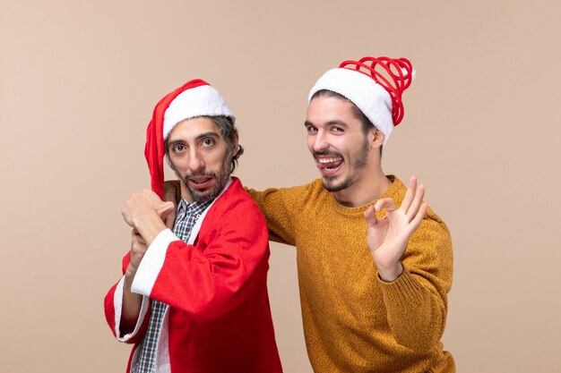 Vorderansicht zwei Freunde mit Weihnachtsmützen, die eine gute Geste auf beigem isoliertem Hintergrund machen