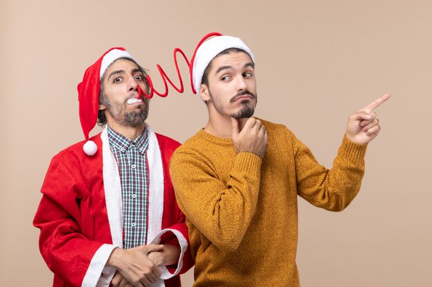Vorderansicht zwei Freunde mit Weihnachtsmützen, die einen Pompon der Fanghüte und die andere Richtung auf beigem isoliertem Hintergrund zeigen