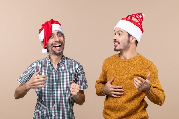 Vorderansicht zwei glückliche Kerle mit Weihnachtsmützen, die auf beigem lokalisiertem Hintergrund lachen