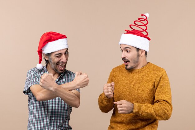 Vorderansicht zwei glückliche Kerle mit Weihnachtsmützen, von denen einer seine Stärke zur Kamera auf beigem isoliertem Hintergrund zeigt