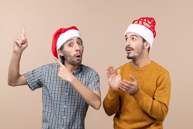 Vorderansicht zwei interessierte Kerle mit Weihnachtsmützen, die etwas dem anderen auf beigem isoliertem Hintergrund zeigen