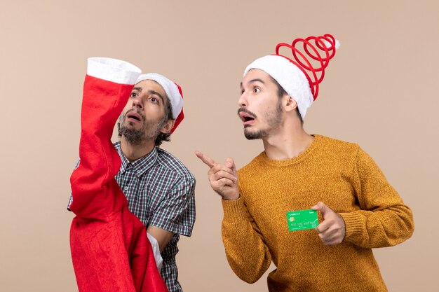 Vorderansicht zwei Kerle, die einen Santa Mantel und den anderen mit Kreditkarte halten, die etwas auf beigem isoliertem Hintergrund betrachten