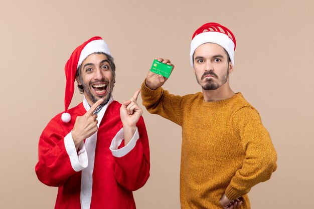 Vorderansicht zwei Kerle einer mit Santa Mantel und der andere mit Kredit, der seine Hand auf eine Taille auf beige lokalisiertem Hintergrund legt