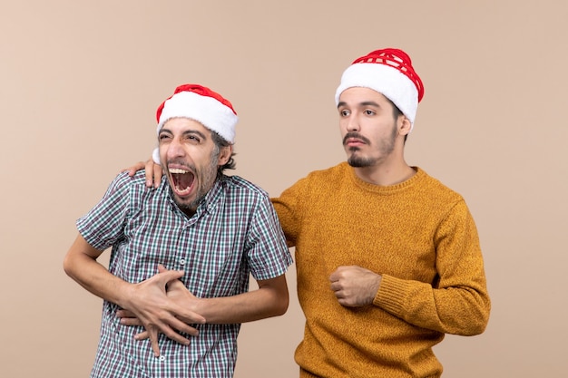 Vorderansicht zwei Kerle mit traurigen Weihnachtsmützen, die seine Hand auf die andere Schulter auf beige lokalisiertem Hintergrund setzen