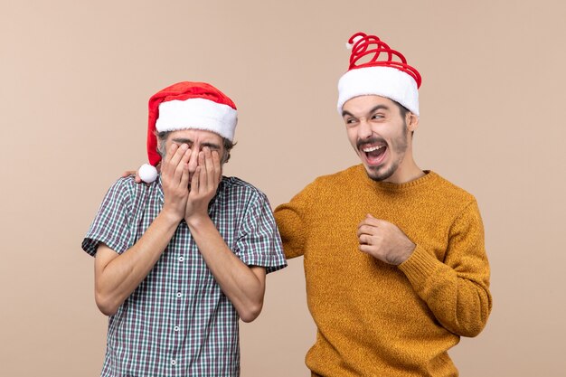 Vorderansicht zwei Kerle mit Weihnachtsmützen, einer bedeckt sein Gesicht mit den Händen und der andere lacht auf beigem isoliertem Hintergrund