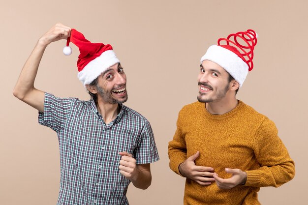 Vorderansicht zwei lachende Kerle mit Weihnachtsmützen, von denen einer seinen Hut auf beigem isoliertem Hintergrund zeigt