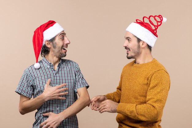 Vorderansicht zwei lächelnde Kerle mit Weihnachtsmützen, die einander auf beige lokalisiertem Hintergrund betrachten